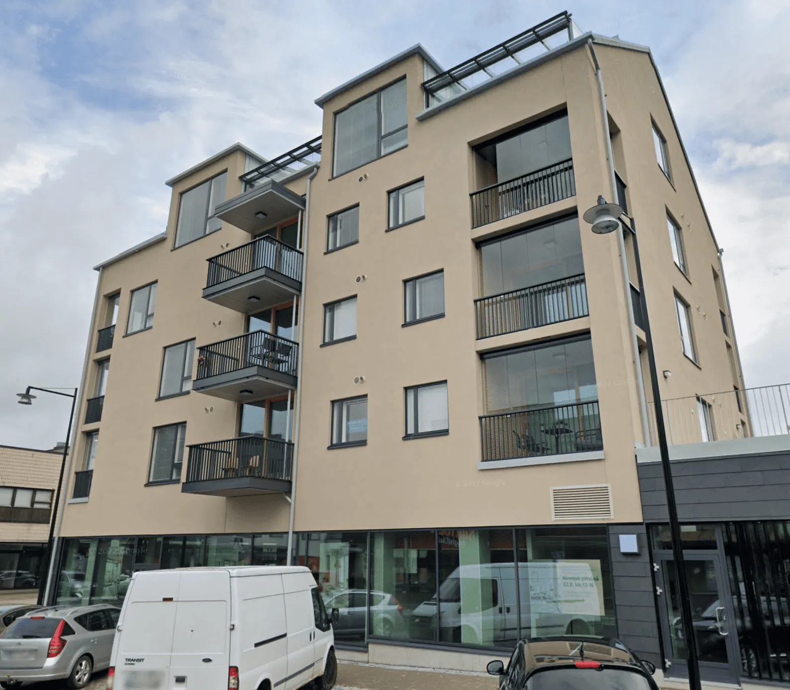 Beige kerrostalo, jossa on parvekkeita, ikkunoita ja autoja edessä pilvisen taivaan alla.